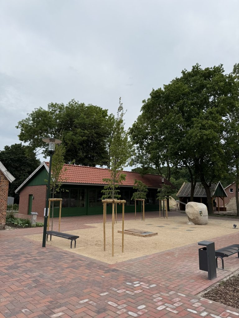 Dorfplatz Lotup, mit Pflasterung, Bäumen und Bänken.